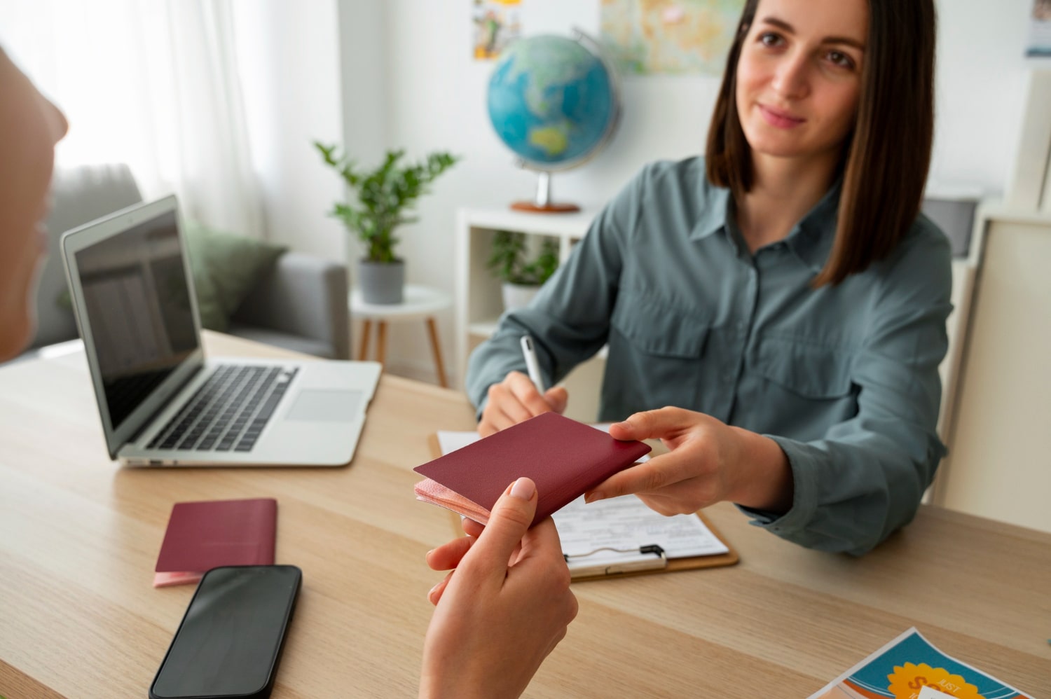 Consultant handing a maroon passport to a client across a desk with documents, laptop, and travel materials during a Cyprus residency consultation.